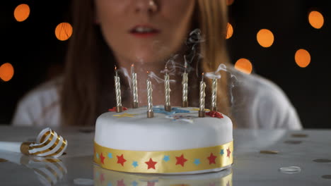 Close-up-of-a-woman-blowing-out-seven-lit-candles-on-a-white,-decorated-birthday-cake,-a-party-blower-beside-it,-bokeh-lights-in-the-background