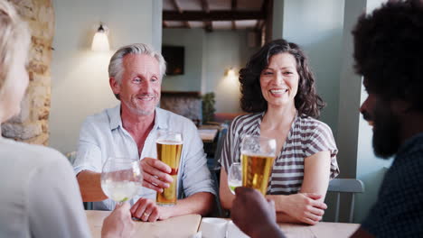 Senior-white-man-and-Hispanic-woman-sitting-at-a-table-in-a-pub-making-a-toast-with-colleagues,-close-up,-over-shoulder-view