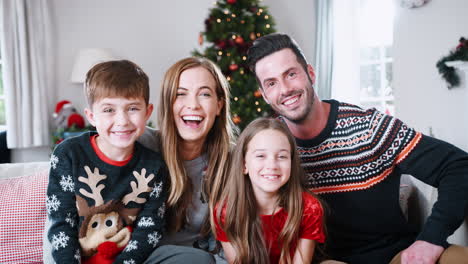 Portrait-Of-Parents-With-Children-Wearing-Festive-Jumpers-Sitting-On-Sofa-In-Lounge-At-Home-On-Christmas-Day