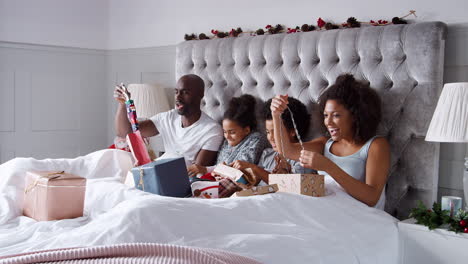 Young-mixed-race-family-sitting-up-in-bed-together-unwrapping-presents-on-Christmas-morning