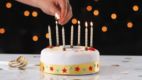 Close-up-of-a-hand-using-a-match-to-light-seven-candles-on-a-white,-decorated-birthday-cake,-a-party-blower-beside-it,-bokeh-lights-in-the-background