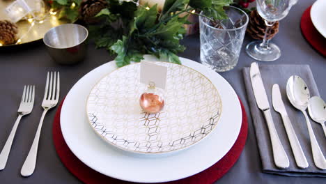 Christmas-table-place-setting-with-bauble-name-card-holder-arranged-on-a-plate-and-green-and-red-table-decorations,-detail,-rack-focus,-elevated-view