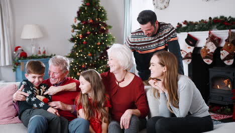 Porträt-Einer-Familie-Aus-Mehreren-Generationen,-Die-Am-Weihnachtstag-Auf-Dem-Sofa-Im-Wohnzimmer-Sitzt-Und-Einen-Festlichen-Pullover-Trägt