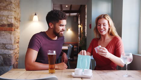 Jump-cut-sequence-of-a-happy-young-blonde-woman-sitting-at-a-table-in-a-pub-receiving-a-gift-from-her-boyfriend,-side-view