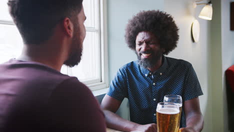 Middle-aged-black-father-having-a-beer-and-talking-with-his-adult-son-sitting-at-table-in-a-pub,-close-up,-dad-facing-camera