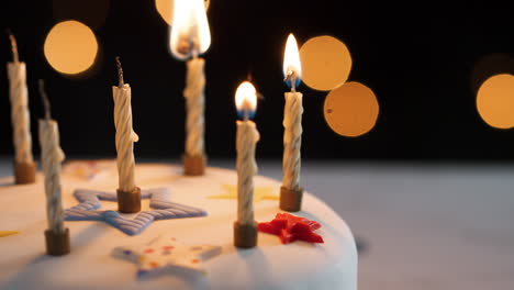 Nahaufnahme-Von-Vier-Kerzen,-Die-Mit-Einem-Streichholz-Angezündet-Werden,-Unter-Unbeleuchteten-Kerzen-Auf-Einem-Weißen,-Dekorierten-Geburtstagskuchen,-Bokeh-Lichter-Im-Hintergrund