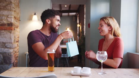 Happy-young-adult-woman-is-surprised-by-a-gift-from-her-boyfriend-while-they-sit-at-table-with-drinks-in-a-pub,-close-up
