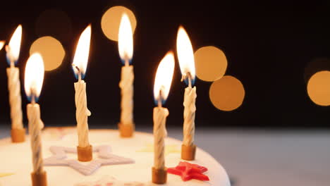 Close-up-of-lit-candles-on-a-white,-decorated-birthday-cake,-bokeh-lights-in-the-background