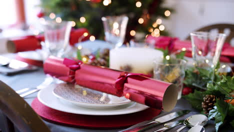 Rack-focus-shot-of-Christmas-dinner-table-with-red-crackers-arranged-on-plates-and-seasonal-decorations,-a-Christmas-tree-with-lights-in-the-background,-selective-focus