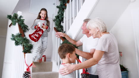 Grandparents-Greeting-Excited-Grandchildren-Wearing-Pajamas-Running-Down-Stairs-Holding-Stockings-On-Christmas-Morning