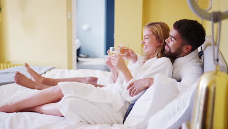 Happy-mixed-race-young-adult-couple-reclining-on-the-bed-in-a-hotel-room-in-bathrobes-holding-glasses-of-wine,-close-up
