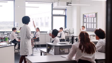 Female-High-School-Tutor-Teaching-Students-In-Uniforms-Sitting-At-Work-Benches-In-Science-Class