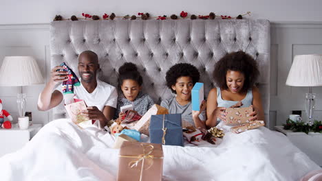 Junge-Gemischtrassige-Familie,-Die-Zusammen-Im-Bett-Sitzt-Und-Am-Weihnachtsmorgen-Geschenke-Auspackt,-Vorderansicht