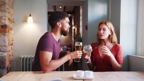Mixed-race-young-adult-couple-sitting-at-table-in-a-pub-holding-drinks-and-talking,-side-view