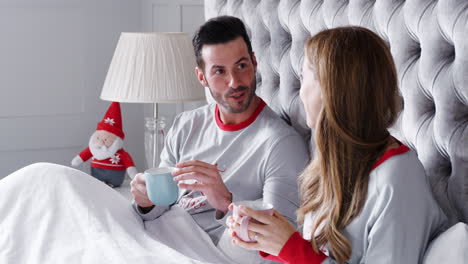 Couple-Wearing-Festive-Pajamas-Relaxing-With-Hot-Drink-In-Bed-At-Christmas