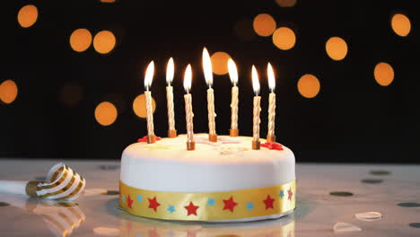 Seven-lit-candles-on-a-white,-decorated-birthday-cake,-a-party-blower-beside-it,-bokeh-lights-in-the-background