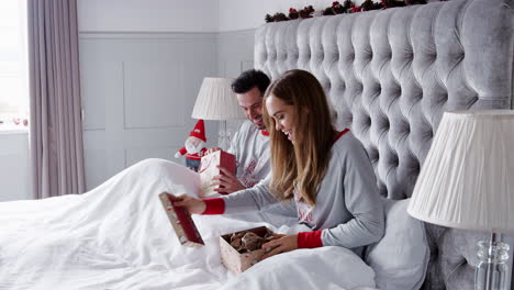 Woman-Opening-Gift-Of-Necklace-In-Bed-At-Home-As-Couple-Exchange-Presents-On-Christmas-Day