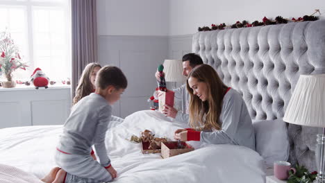 Parents-Opening-Gifts-From-Children-As-They-Sit-On-Bed-Exchanging-Present-On-Christmas-Day