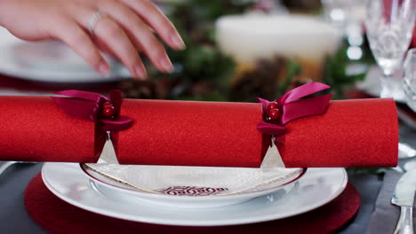 A-woman’s-hand-laying-a-red-Christmas-cracker-on-a-plate-at-a-table-decorated-for-Christmas-dinner,-close-up
