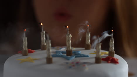 Close-up-of-a-woman-blowing-out-seven-lit-candles-on-a-white,-decorated-birthday-cake,-a-party-blower-beside-it,-bokeh-lights-in-the-background,-detail
