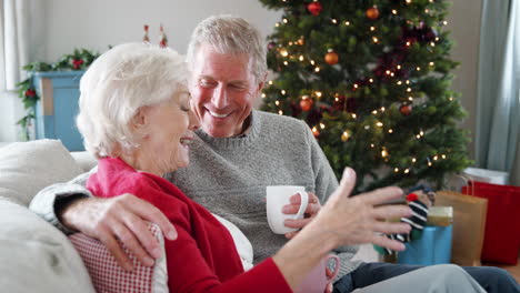 Close-Up-Of-Senior-Couple-Sitting-On-Sofa-With-Hot-Drink-At-Home-And-Chatting-With-Christmas-Tree-In-Background