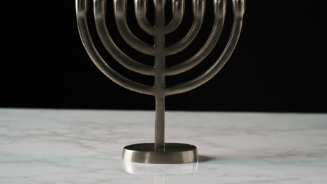 Tilt-shot-of-Jewish-menorah-candelabrum-with-lit-candles-on-a-grey-marble-surface,-against-black-background
