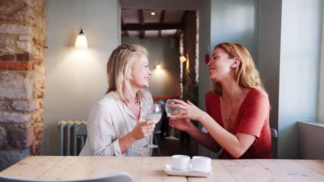 Two-young-adult-blonde-women-holding-wine-glasses-talking-with-each-other-sitting-at-a-table-in-a-pub,-close-up,-side-view