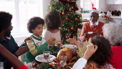 Mixed-race,-multi-generation-family-sitting-at-their-Christmas-dinner-table-reading-jokes-and-putting-on-paper-party-hats-from-Christmas-crackers,-close-up