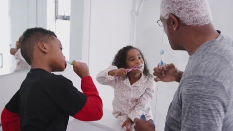 Grandfather-In-Bathroom-Wearing-Pajamas-Helping-Grandchildren-To-Brush-Teeth