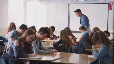 Male-High-School-Teacher-Walking-Between-Desks-As-Students-Work-During-Lesson
