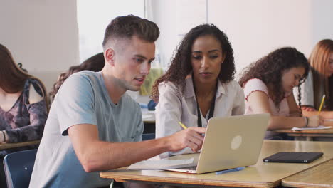 Female-Tutor-Giving-One-To-One-Support-To-Male-Student-Working-At-Desk-On-Laptop