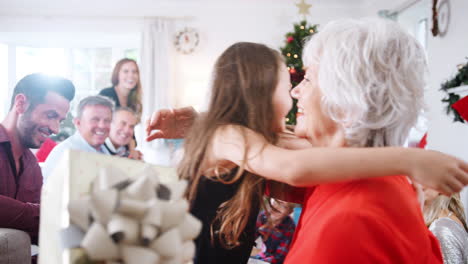 Enkelin-Gibt-Der-Großmutter-Ein-Geschenk,-Während-Die-Familie-Aus-Drei-Generationen-In-Der-Lounge-Sitzt-Und-Gemeinsam-Den-Weihnachtstag-Zu-Hause-Feiert