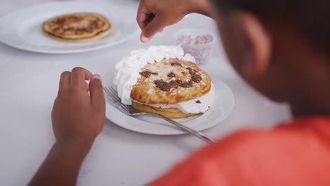 Close-Up-Of-Boy-Adding-Sprinkles-To-Pancakes-With-Face-Made-From-Cream-And-Chocolate-Sauce-In-Kitchen