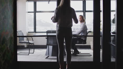 Young-white-woman-joins-male-creative-in-a-meeting-room-for-a-job-interview,-seen-through-glass-wall