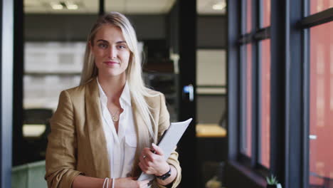Millennial-white-blonde-businesswoman-holding-documents,-turns-and-smiles-to-camera,-close-up