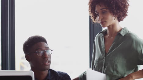 Millennial-black-woman-stands-talking-to-her-male-creative-colleague-at-his-desk,-close-up