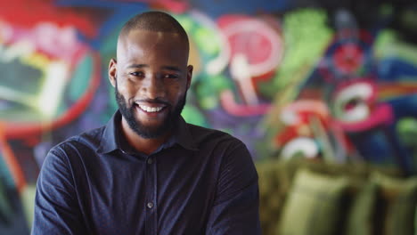 Millennial-black-male-creative-in-front-of-mural-in-workplace-smiling-to-camera,-head-and-shoulders