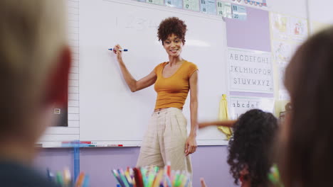Female-Teacher-Standing-At-Whiteboard-Teaching-Lesson-To-Elementary-Pupils-In-School-Classroom