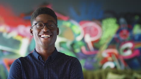 Young-black-male-creative-smiling-to-camera-in-front-of-mural-in-casual-office,-head-and-shoulders