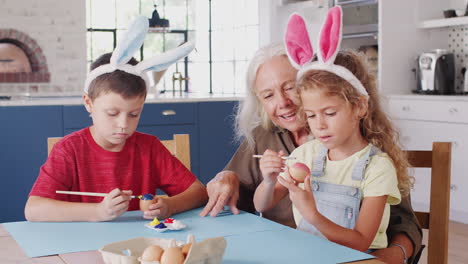 Abuela-Con-Nietos-Usando-Orejas-De-Conejo-Decorando-Huevos-De-Pascua-Juntos-En-Casa
