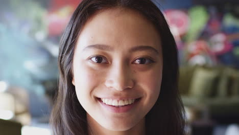 Young-Asian-woman-smiling-to-camera-in-front-of-mural-at-creative-workplace,-head-shot,-close-up