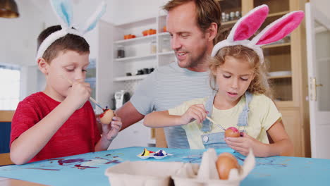 Father-With-Children-Wearing-Rabbit-Ears-Decorating-Easter-Eggs-At-Home-Together