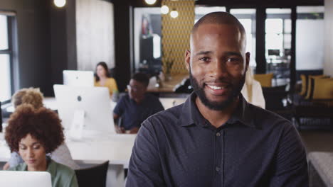 Bearded-millennial-black-male-creative-standing-in-a-busy-casual-office,-smiling-to-camera,-close-up