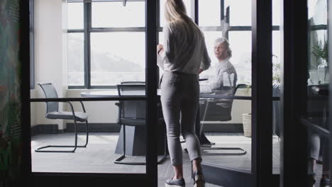 Young-white-woman-joins-male-creative-in-a-meeting-room-for-a-job-interview,-seen-through-glass-wall