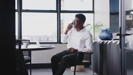Millennial-black-businessman-in-an-office-answers-his-phone,-puts-his-feet-up-on-the-table,-close-up