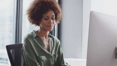 Millennial-black-woman-sitting-at-an-office-desk-using-a-computer-and-drinking-coffee,-waist-up