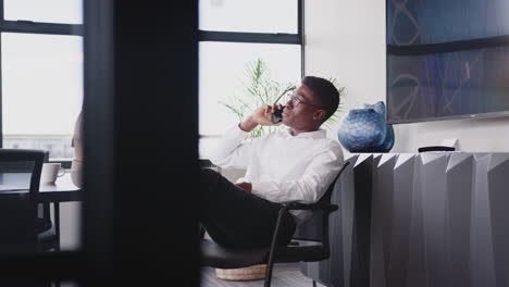 Young-black-man-talking-on-phone-in-an-office,-feet-up-on-table,-close-up,-seen-through-glass-wall