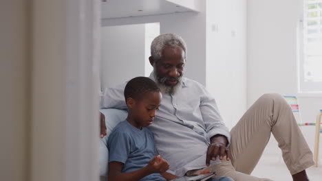 Grandfather-Sitting-With-Grandson-In-Childs-Bedroom-Using-Digital-Tablet-Together
