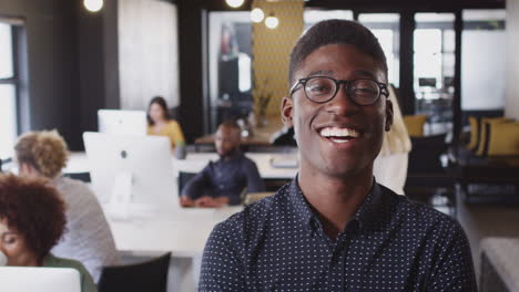 Millennial-black-male-creative-wearing-glasses-smiling-to-camera-in-a-busy-casual-office,-close-up