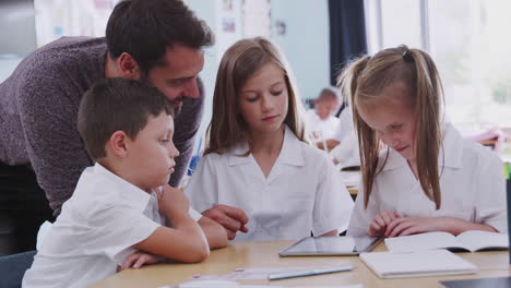 Male-Teacher-With-Three-Elementary-School-Pupils-Wearing-Uniform-Using-Digital-Tablet-At-Desk